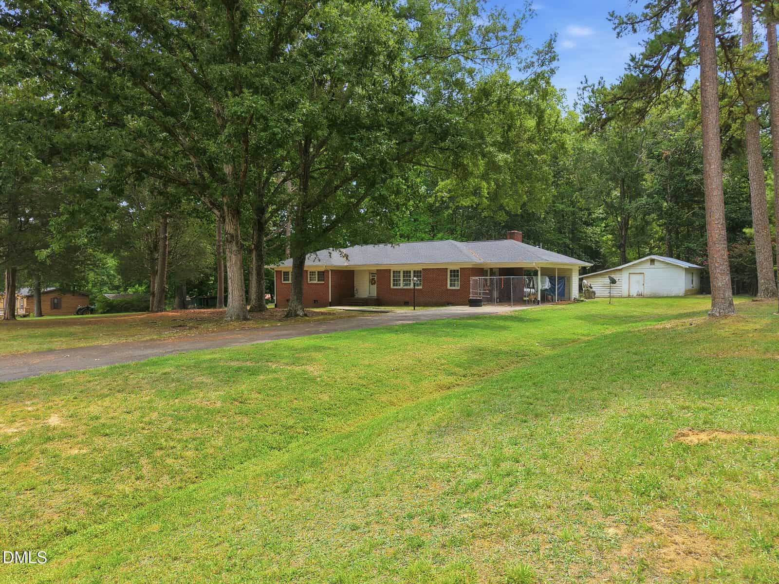 Single-story brick house with a covered carport, large trees in front yard, spacious grassy lawn, and a separate white garage in the background.