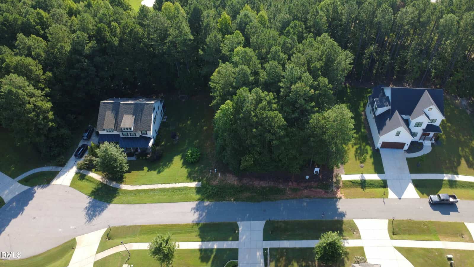 Aerial view of two houses separated by a wooded lot, curved roads, and driveways; dense trees fill the middle, while lawns and a parked car are visible on the street.