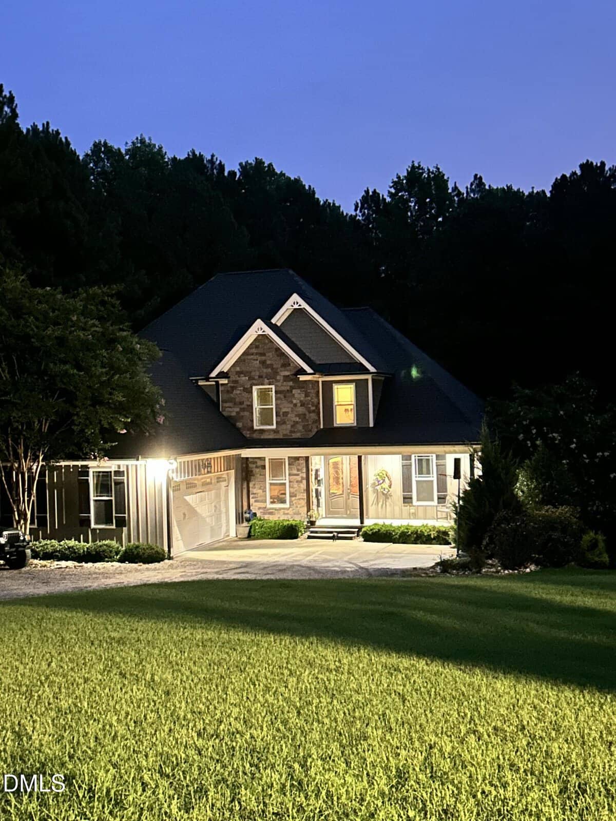 A two-story house with illuminated porch lights sits on a well-manicured lawn at dusk in Wake Forest, surrounded by trees and greenery, with a driveway leading to the garage on the left at 4002 Stoneway Court.