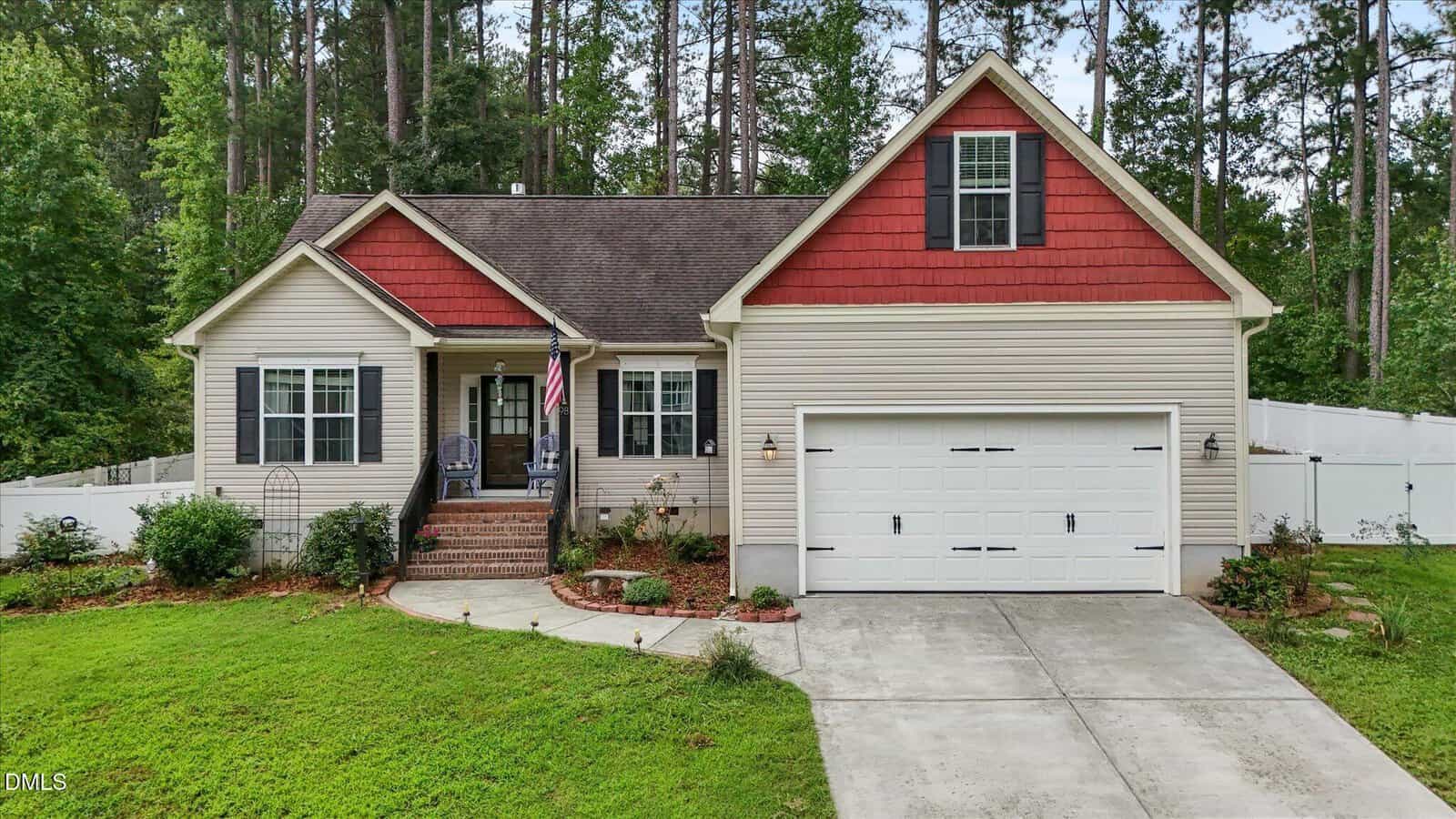Single-story house at 98 Bluegrass Drive, Oxford, featuring beige siding, red gables, a welcoming front porch with chairs and an American flag, an attached two-car garage, classic brick steps, a beautifully landscaped yard, and tall trees in the background.