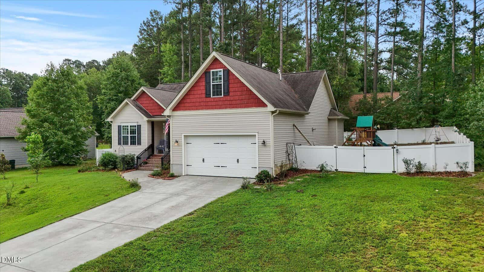 A single-story house with a red gable, attached two-car garage, fenced backyard with playset, and a long driveway, surrounded by grass and tall trees.