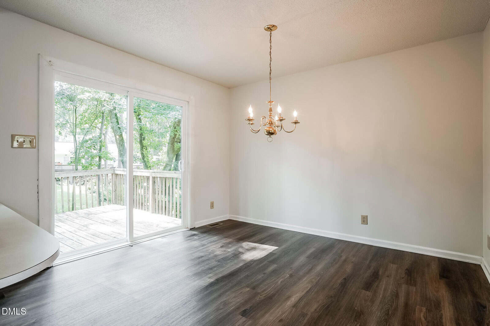 Empty dining room with white walls, wood floors, a gold chandelier, and a sliding glass door leading to a wooden deck outside with trees visible.