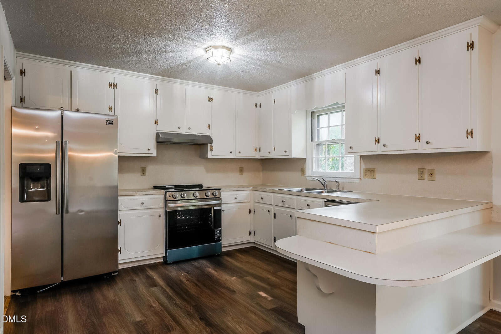 Bright kitchen with white cabinets, stainless steel refrigerator, gas stove, and double sink under a window. The room features laminate wood flooring and a peninsula counter with an overhang for seating.