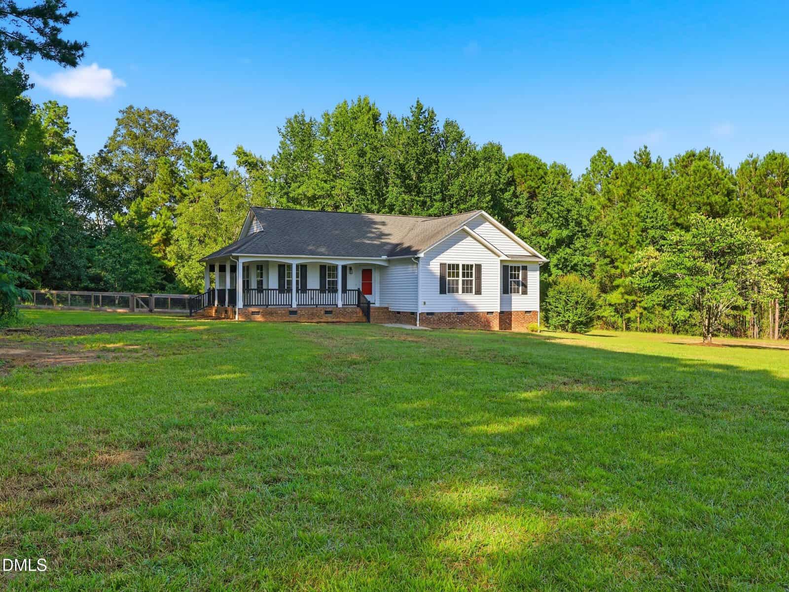 A white one-story house with a porch sits on a large, grassy lawn surrounded by trees under a clear blue sky at 665 Lewis Road in Zebulon.