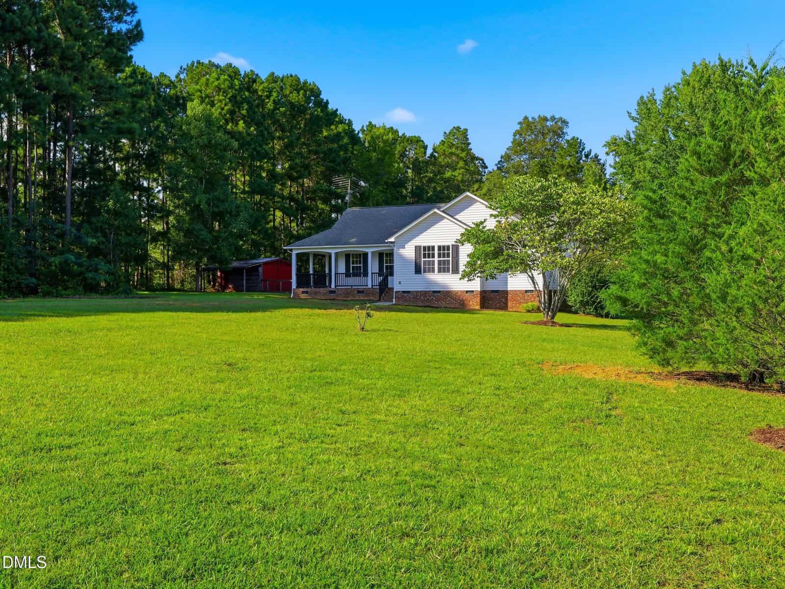 A white house with a front porch sits on a large, green lawn surrounded by trees under a blue sky. A red barn is visible in the background on the left.