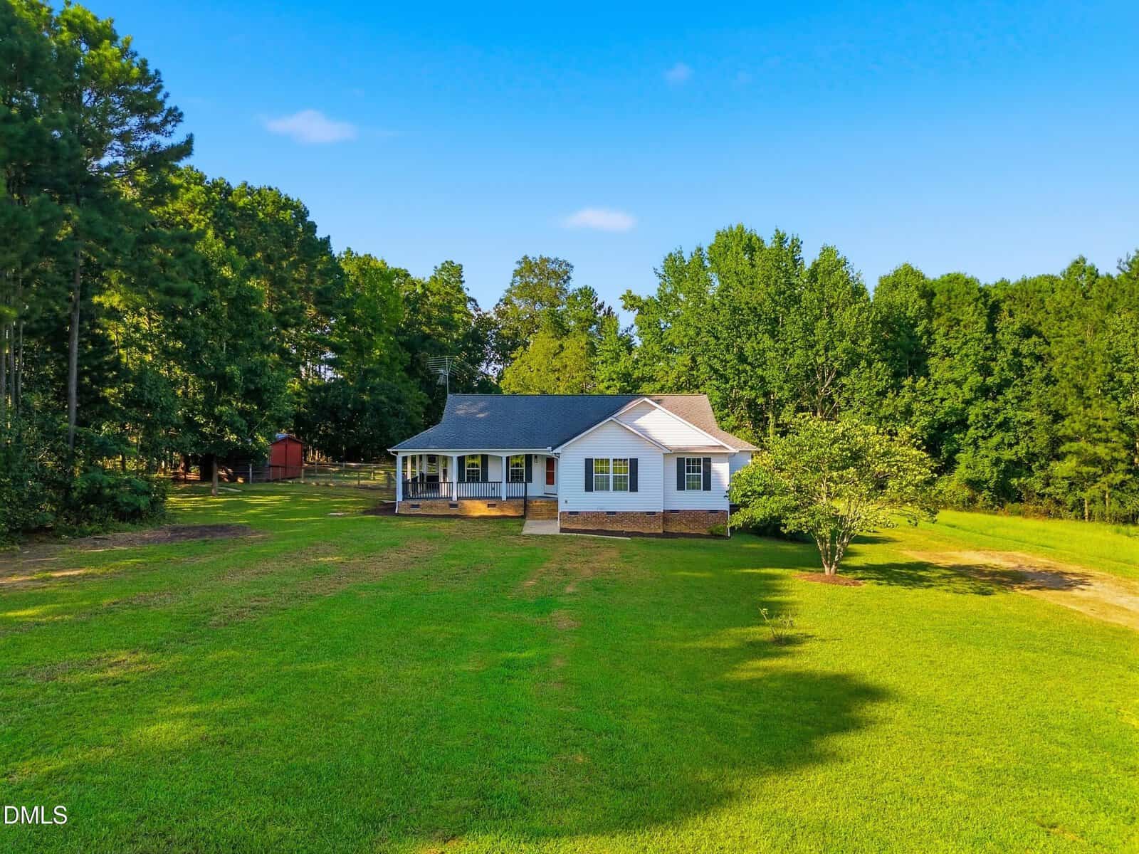 White single-story house with a covered front porch sits on a large, grassy yard surrounded by trees under a clear blue sky.