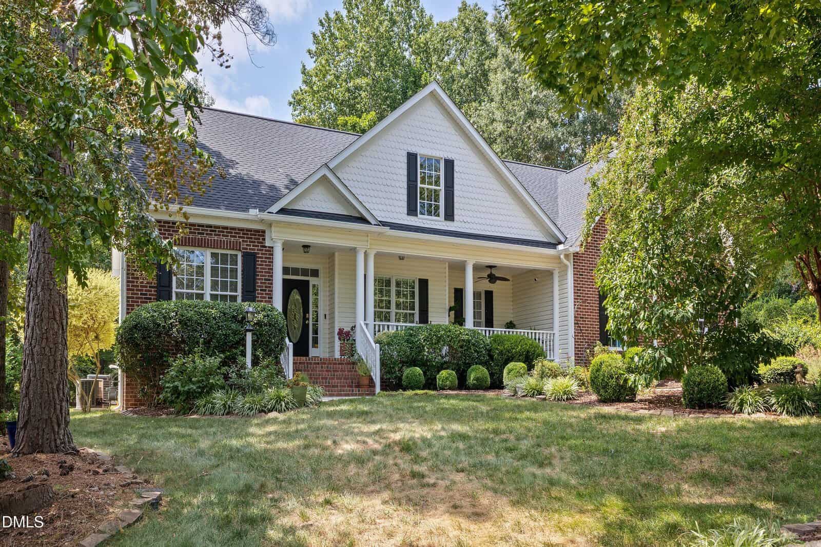 A single-story brick house with white siding, front porch, columns, black shutters, manicured lawn, shrubs, and trees surrounding the yard on a sunny day in the desirable Youngsville community.