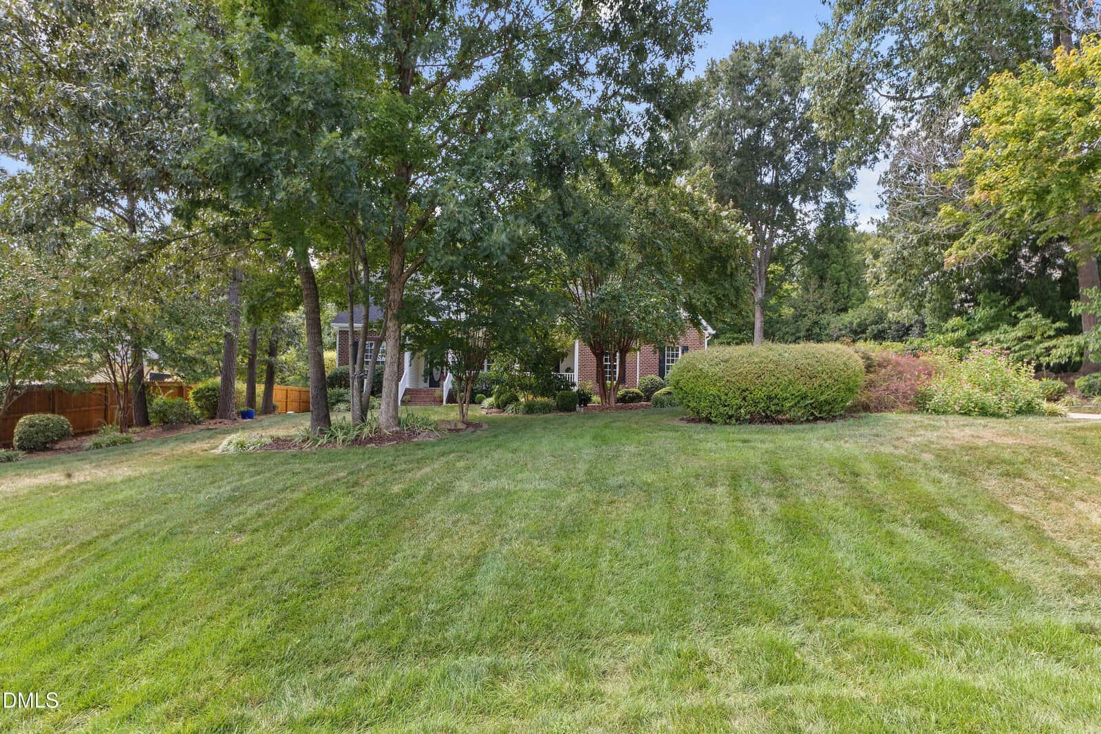 A well-maintained lawn with trees, shrubs, and a brick house partially visible in the background on a sunny day.