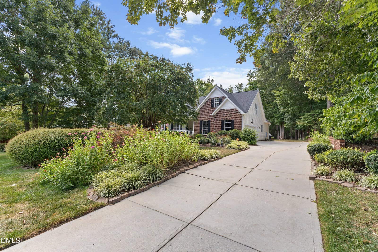 A long driveway leads to a two-story brick and siding house surrounded by lush landscaping, green trees, and well-maintained shrubs on a sunny day.