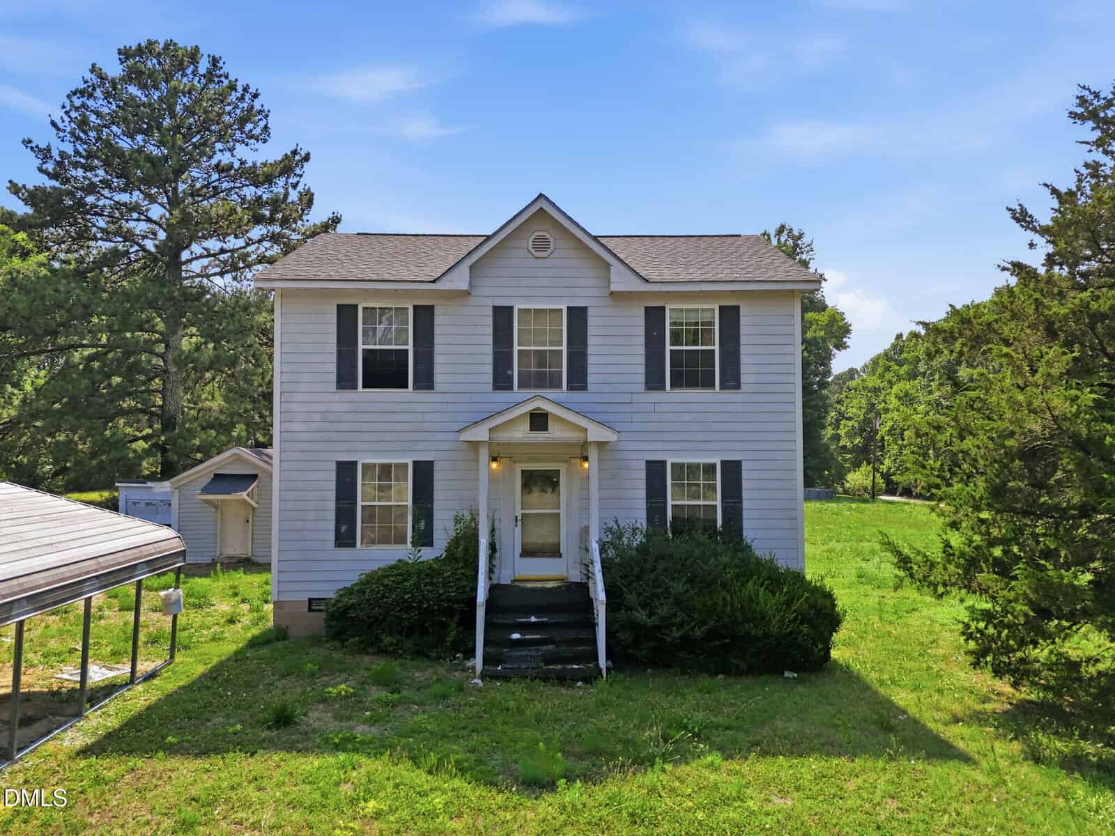 Two-story white house with black shutters, front porch, and overgrown bushes situated at 531 Ballard Pruitt Road in Franklinton. A large grassy yard surrounds the home, with tall trees in the background and a carport visible on the left side. Sky is clear and blue.