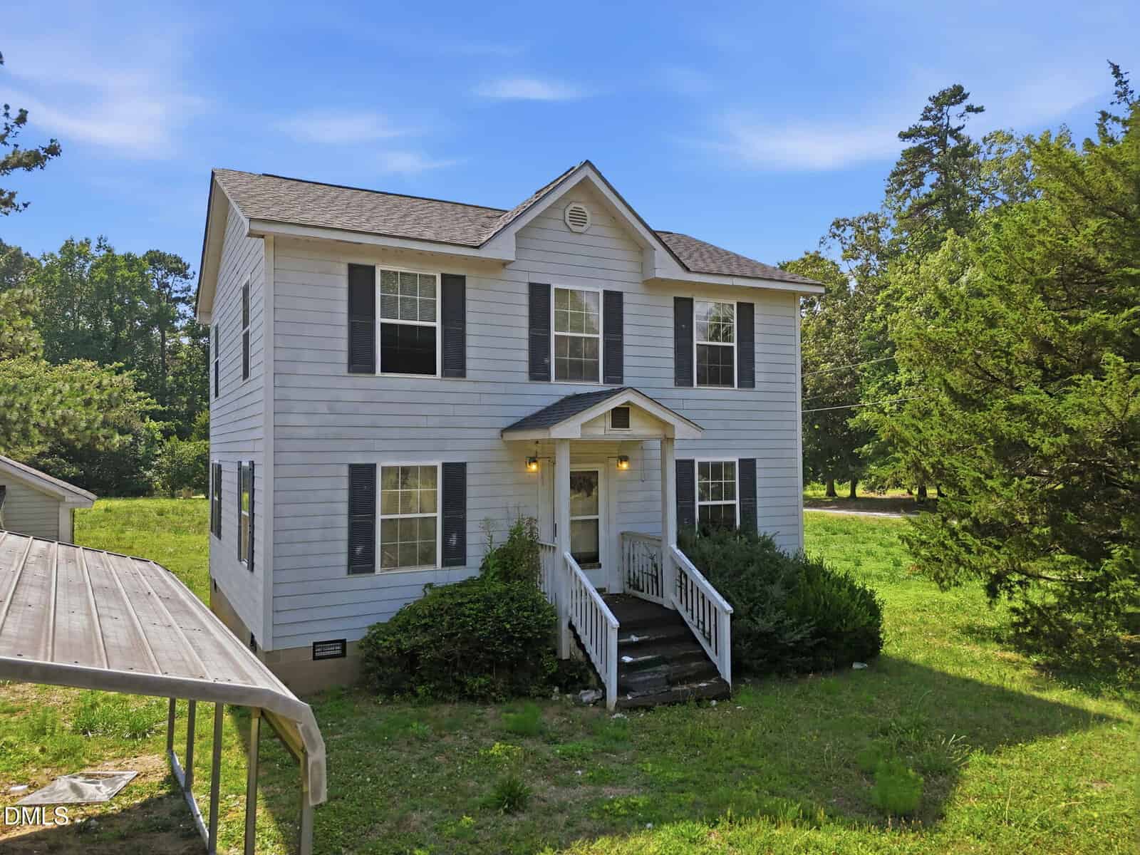 Two-story white house with black shutters, front porch, and steps surrounded by green bushes and grass. A carport is partially visible on the left. Trees and blue sky are in the background.