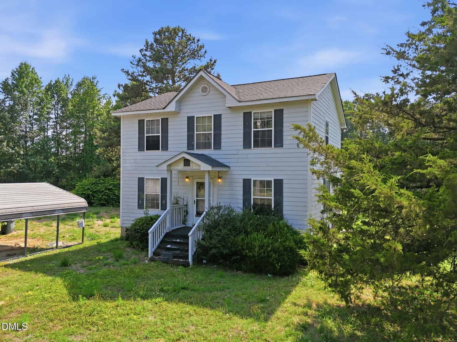 Two-story white house with gray shutters and a covered front porch, surrounded by trees and greenery. A carport is visible to the left under a blue sky.
