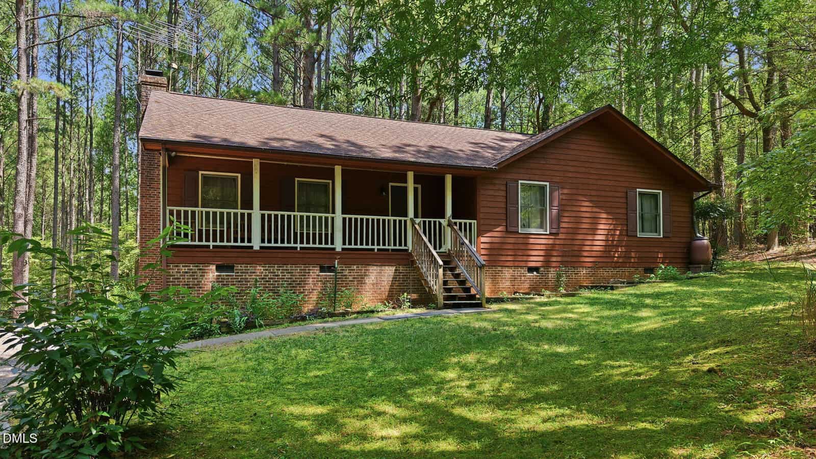 A rustic brown house with a front porch sits on a grassy lawn, surrounded by tall trees. Steps lead up to the porch, and sunlight filters through the forested background.