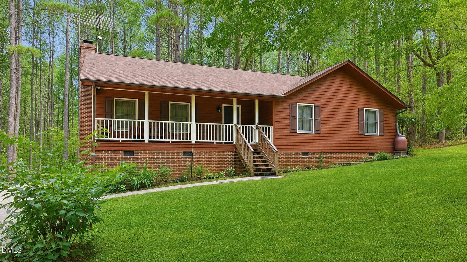 A brown wooden house with a covered front porch, white railings, and steps sits gracefully at 170 River View Drive in Franklinton, surrounded by green grass and tall trees in a tranquil wooded setting.