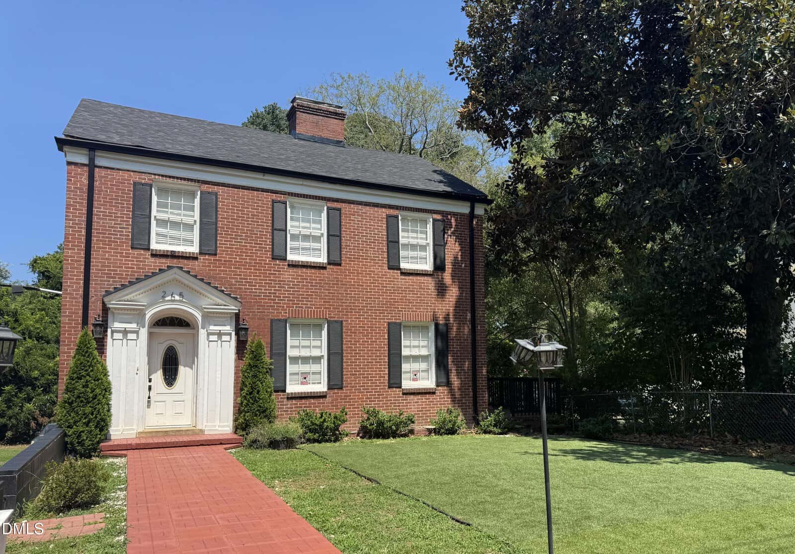 The charming two-story red brick house at 265 Gholson Avenue in Henderson features black shutters, white trim, a graceful white arched front door, and a manicured lawn. Large trees surround the yard, and a black fence is visible on the right side.