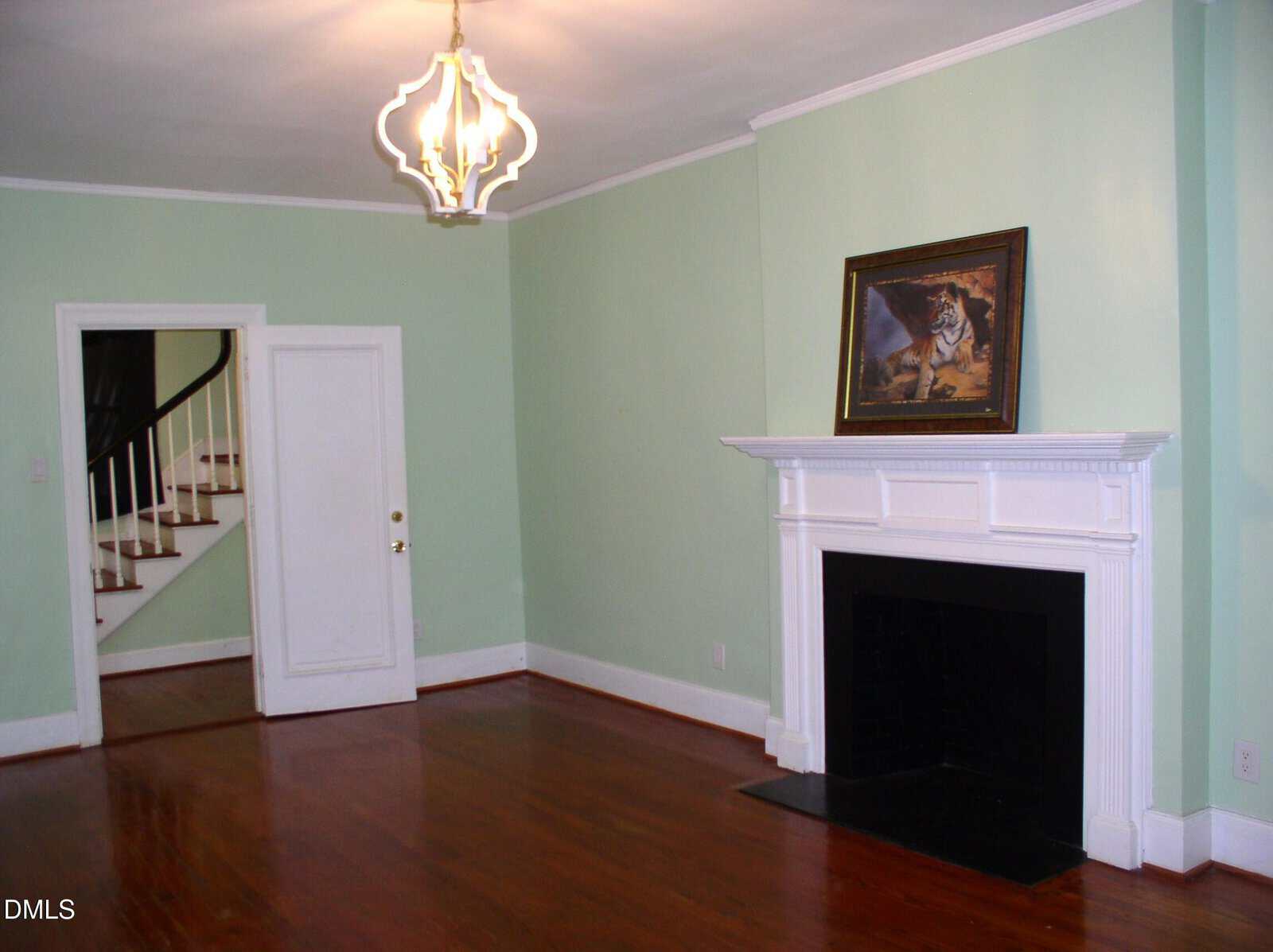 A living room with mint green walls, wooden floor, white fireplace, and a framed tiger painting on the mantel. A white door and open doorway reveal a staircase in the background. Unique modern ceiling light fixture.