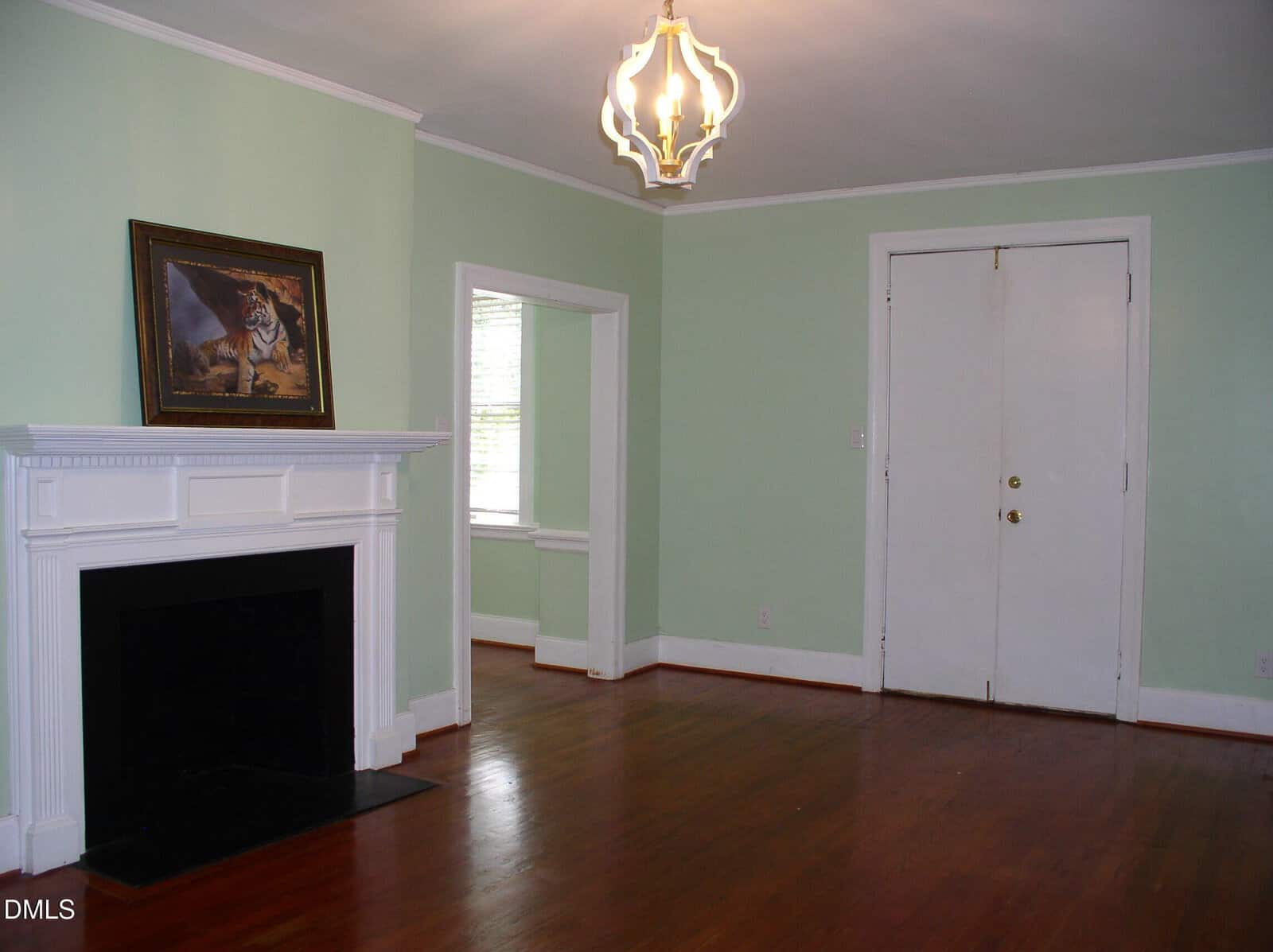 A living room with light green walls, wood floors, a white fireplace with a painting above it, a window with blinds, and double white doors. A modern gold light fixture hangs from the ceiling.