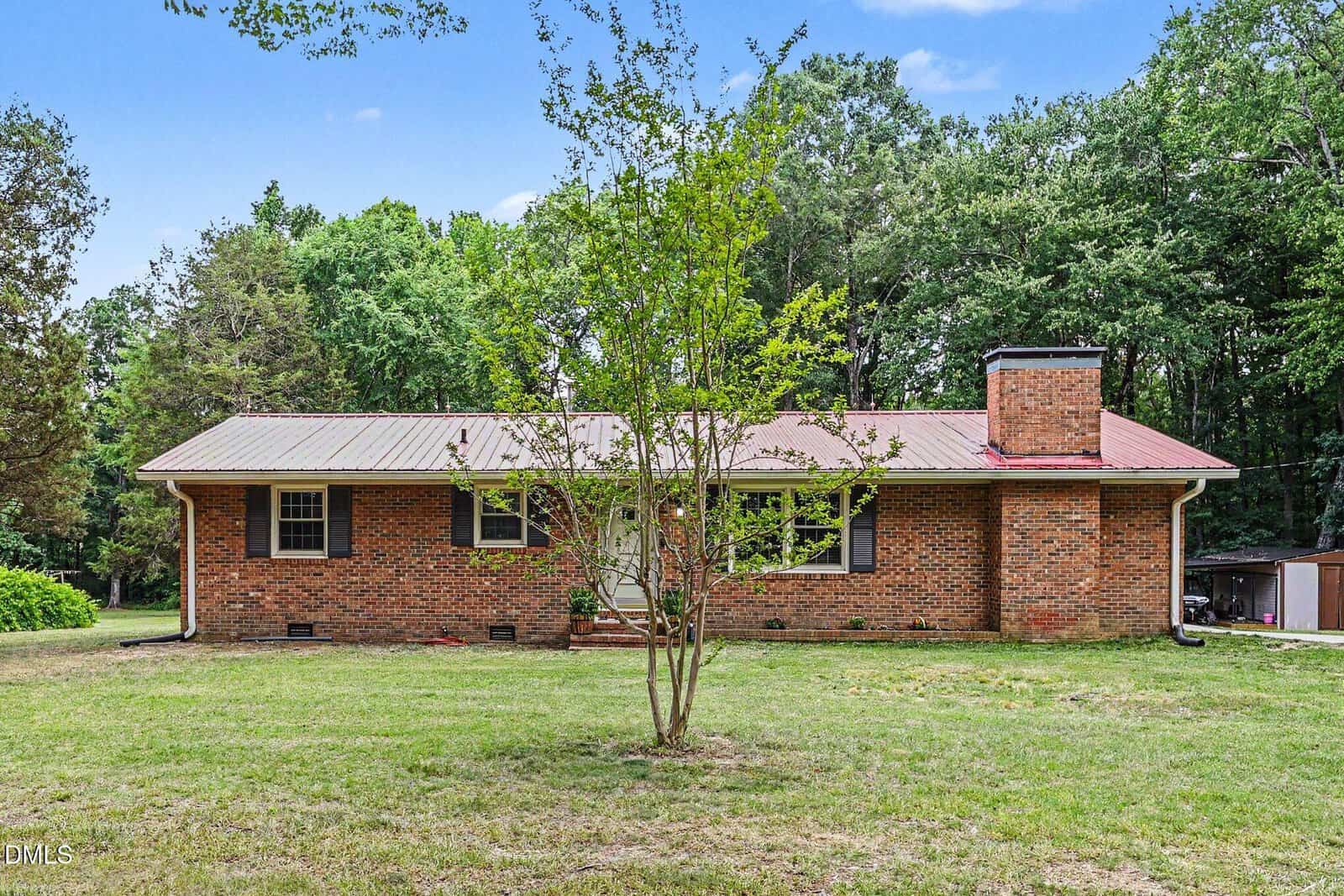 Single-story brick house with a metal roof, large front yard, one front-facing window on each side of the door, a prominent chimney on the right, and surrounded by trees and green grass under a partly cloudy sky—located at 2064 Oak Hill Road in charming Oxford.