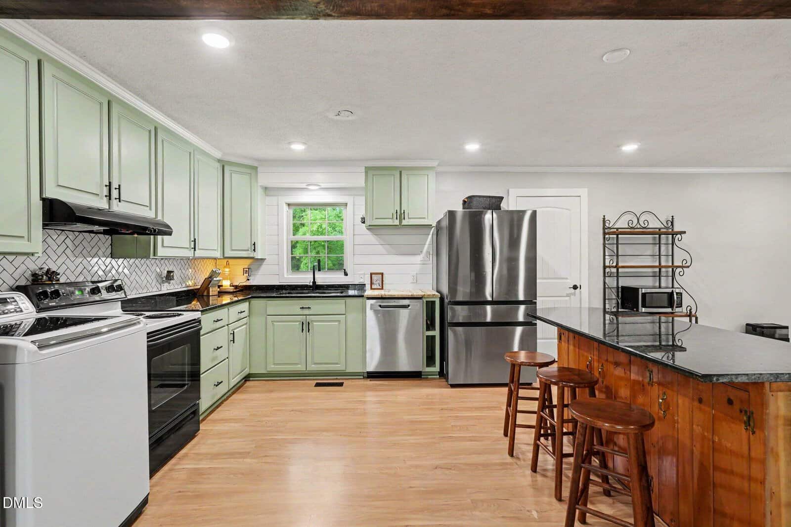 Modern kitchen with light green cabinets, stainless steel appliances, a wooden island with stools, black countertops, a window above the sink, light wood flooring, and a metal shelving unit with a microwave on the right.