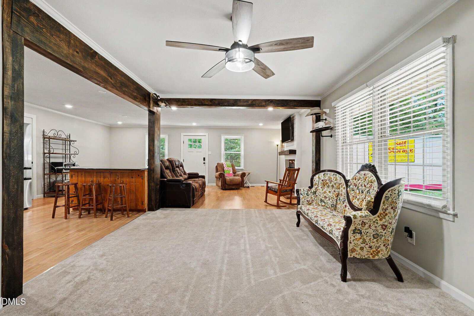 Living room with beige carpet, floral sofa, large windows with blinds, and ceiling fan. Adjacent area has wood flooring, recliners, a bar, and French doors leading outside. Wooden beams separate the spaces.