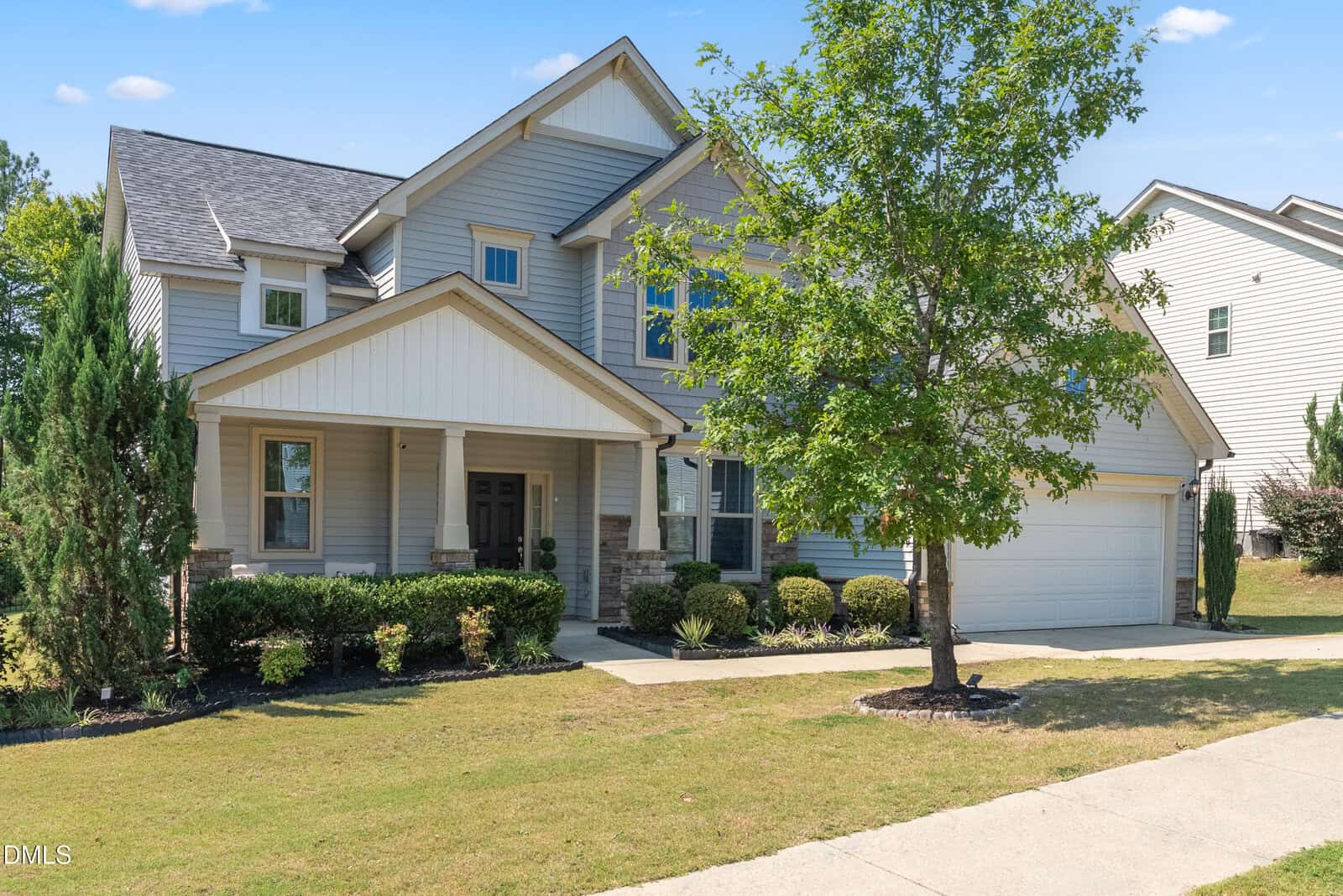 Two-story suburban house with light gray siding, white trim, covered porch, and attached two-car garage. Manicured lawn, shrubs, and a tree in front. Bright, sunny day with a clear blue sky.