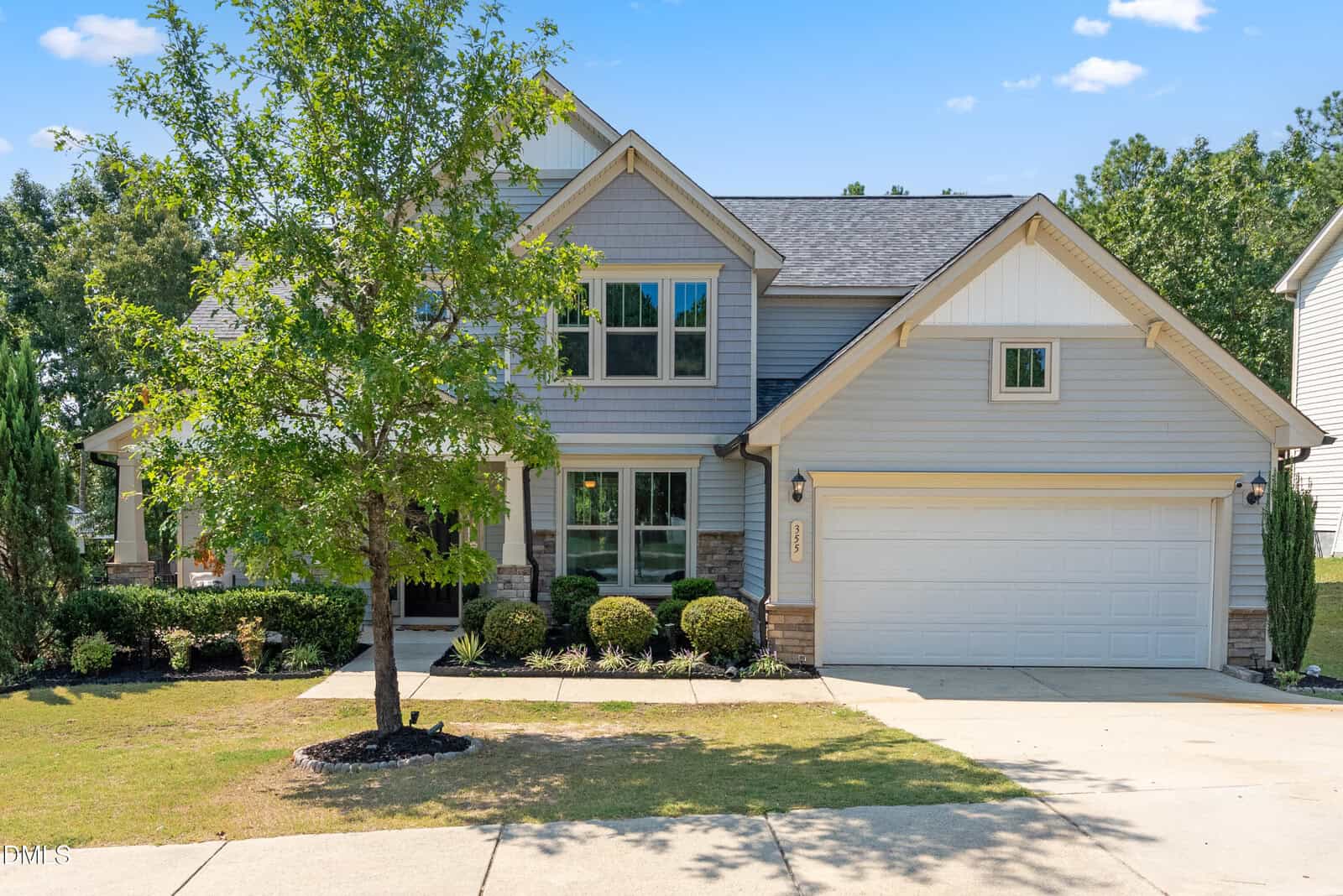 Two-story suburban house with gray siding, white trim, and a two-car garage. There’s a small tree and bushes in the front yard, with a driveway and sidewalk leading to the entrance. Blue sky in the background.