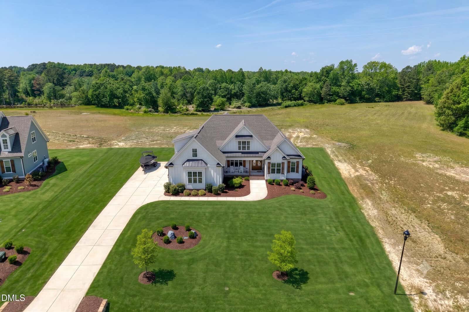 A large, two-story house with a neatly landscaped lawn, driveway, and surrounding fields, set against a backdrop of trees under a clear blue sky.