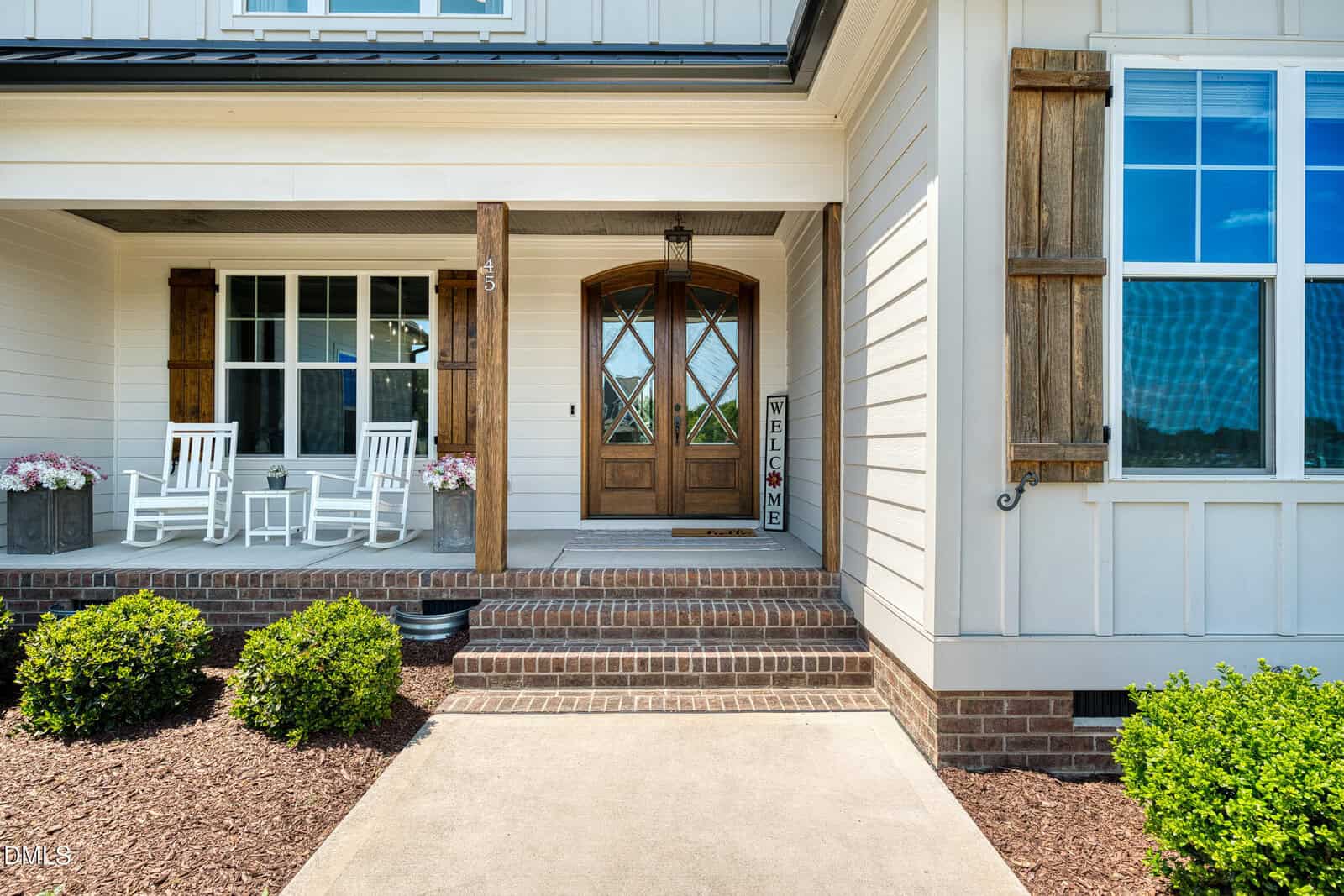 A front porch with two white rocking chairs, a wooden door with glass panels, a welcome sign, brick steps, and shrubs in front of a white house with wooden shutters.
