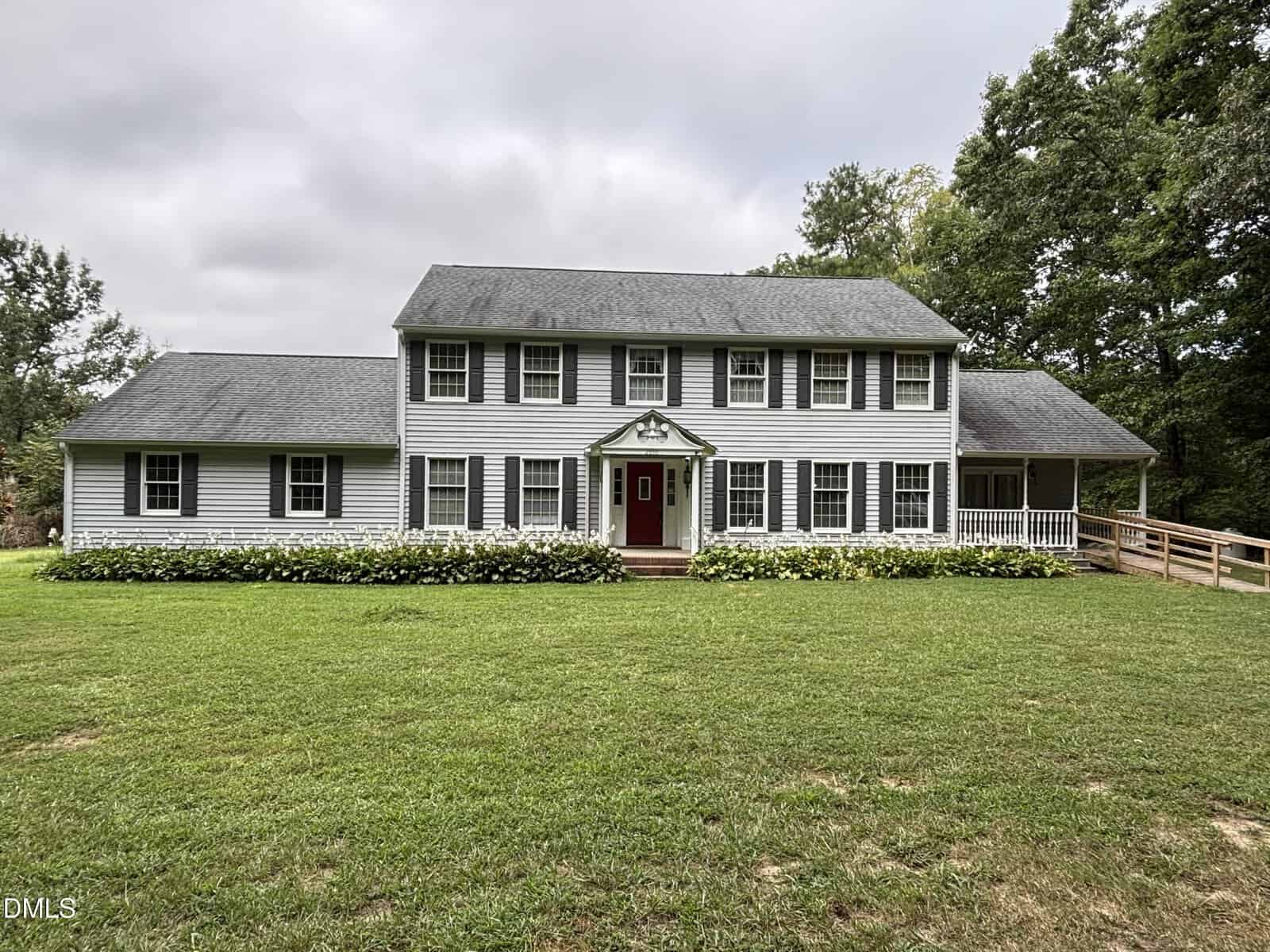 Two-story white house with dark shutters, a red door, columned entrance, and a wide front lawn at 2950 Old Watkins Road. There is a ramp on the right side leading to a covered porch. Trees and bushes surround the building in this charming Henderson property.