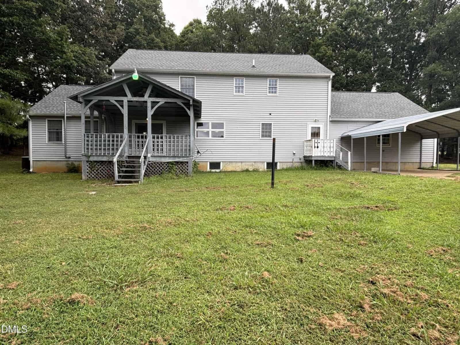 Back view of a two-story gray house with a covered porch, stairs leading to the yard, and an attached carport on the right. The house is surrounded by grass and trees.