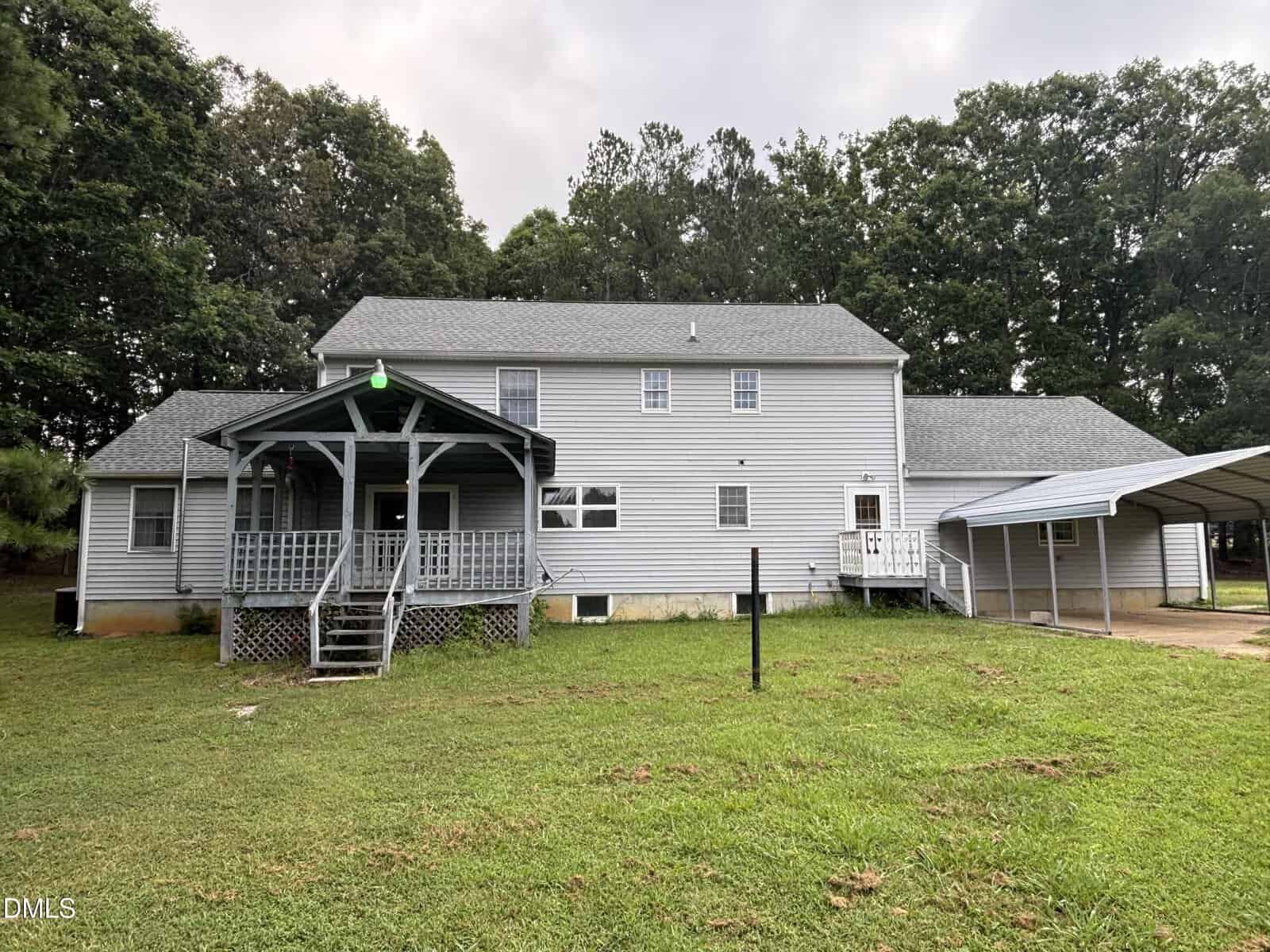 Two-story gray house with a covered front porch, white trim, and a carport on the right, set on a grassy lawn with trees in the background under a cloudy sky.