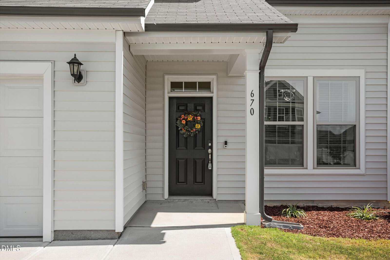 A black front door with a floral wreath, house number 670, and white siding. A window with closed blinds is to the right, and a small garden with mulch is nearby.