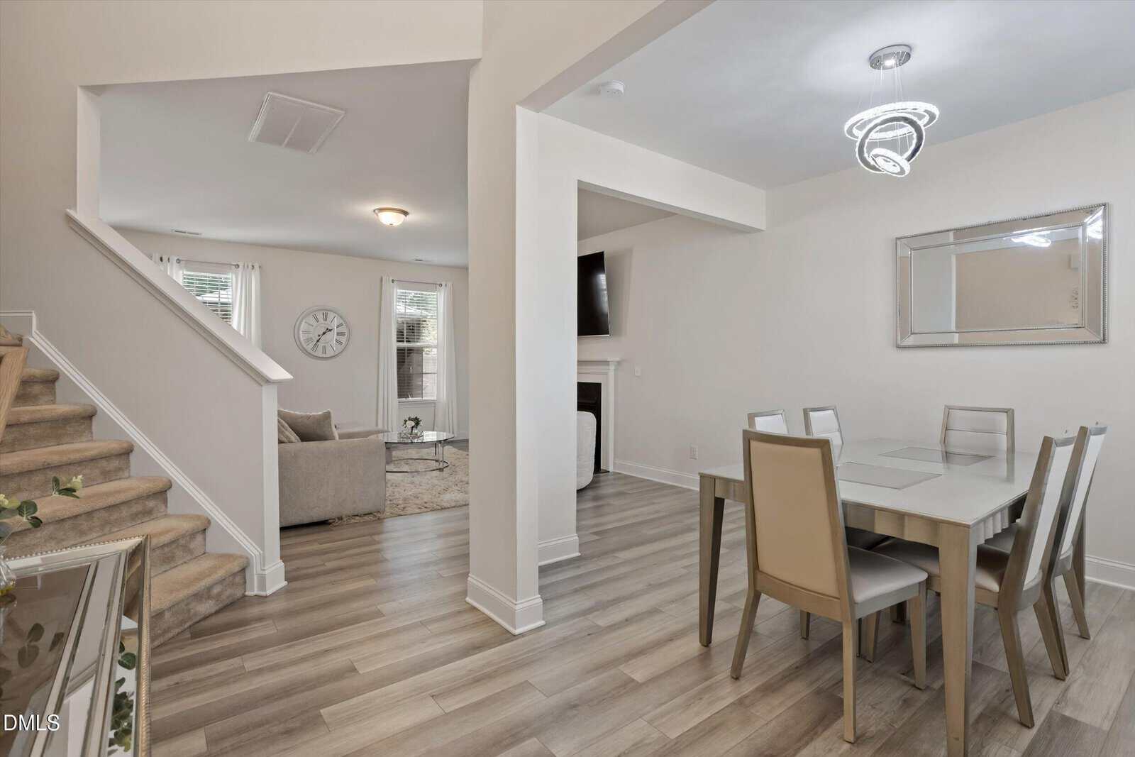 Modern open-concept dining and living area with light wood floors, a beige dining set for six, neutral walls, stairs on the left, and a cozy living area with large windows, beige seating, and a wall clock.