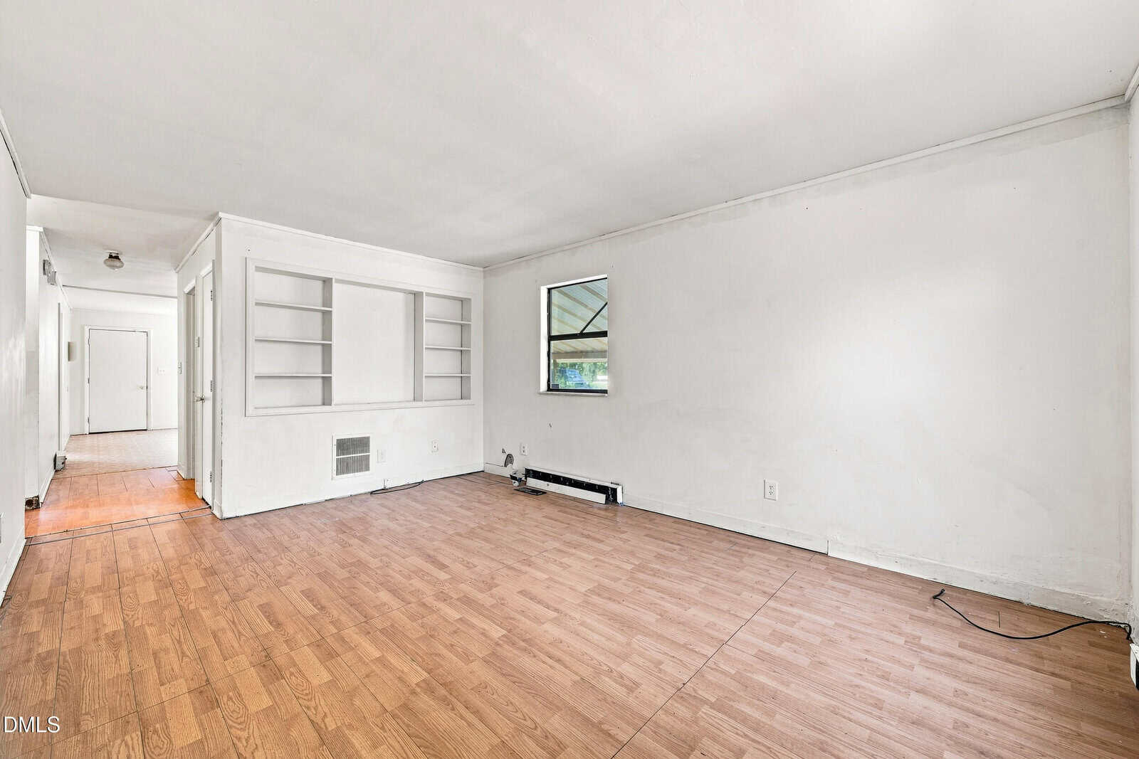 Unfurnished room with light wood flooring, white walls, a single small window, built-in shelves, and a wall heater. Hallway and doors are visible in the background. Room appears clean but minimalistic.