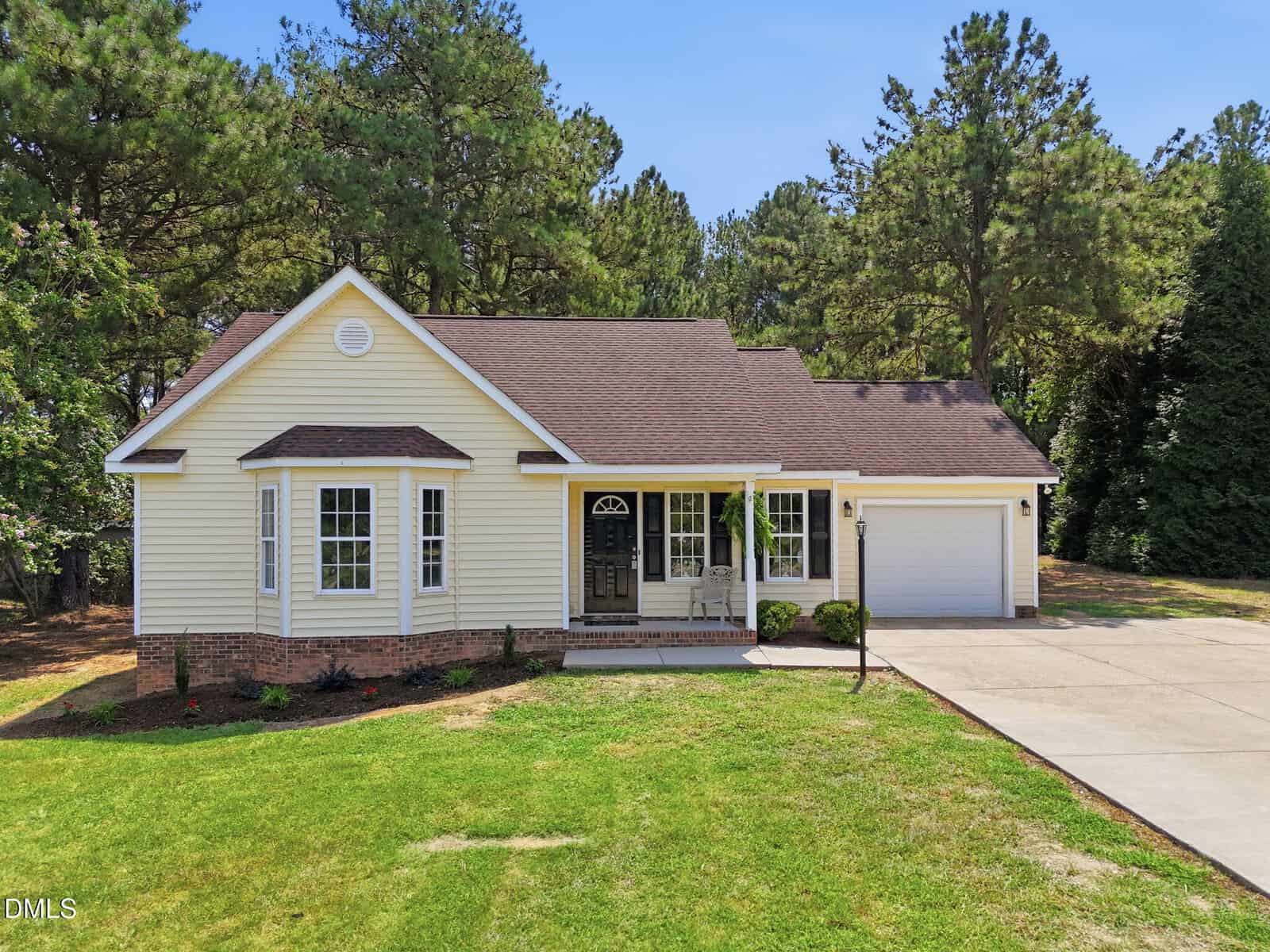 Single-story beige house with a front porch, white trim, and attached two-car garage, surrounded by grass and trees under a clear blue sky. A paved driveway leads up to the garage and front entrance.
