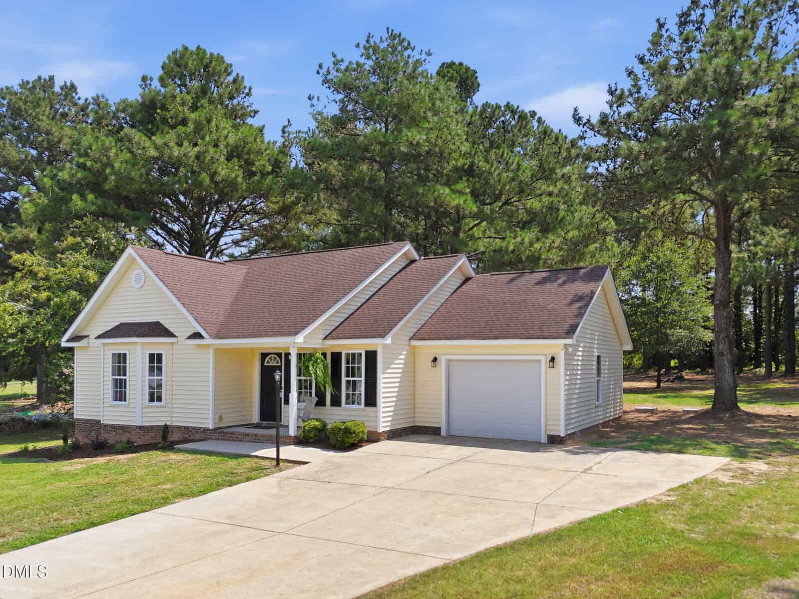 Charming single-story yellow house located at 130 Stillmeadow Drive, Louisburg, with a brown roof, attached one-car garage, and wide driveway. The home is surrounded by green grass, tall pine trees, and a bright blue sky.