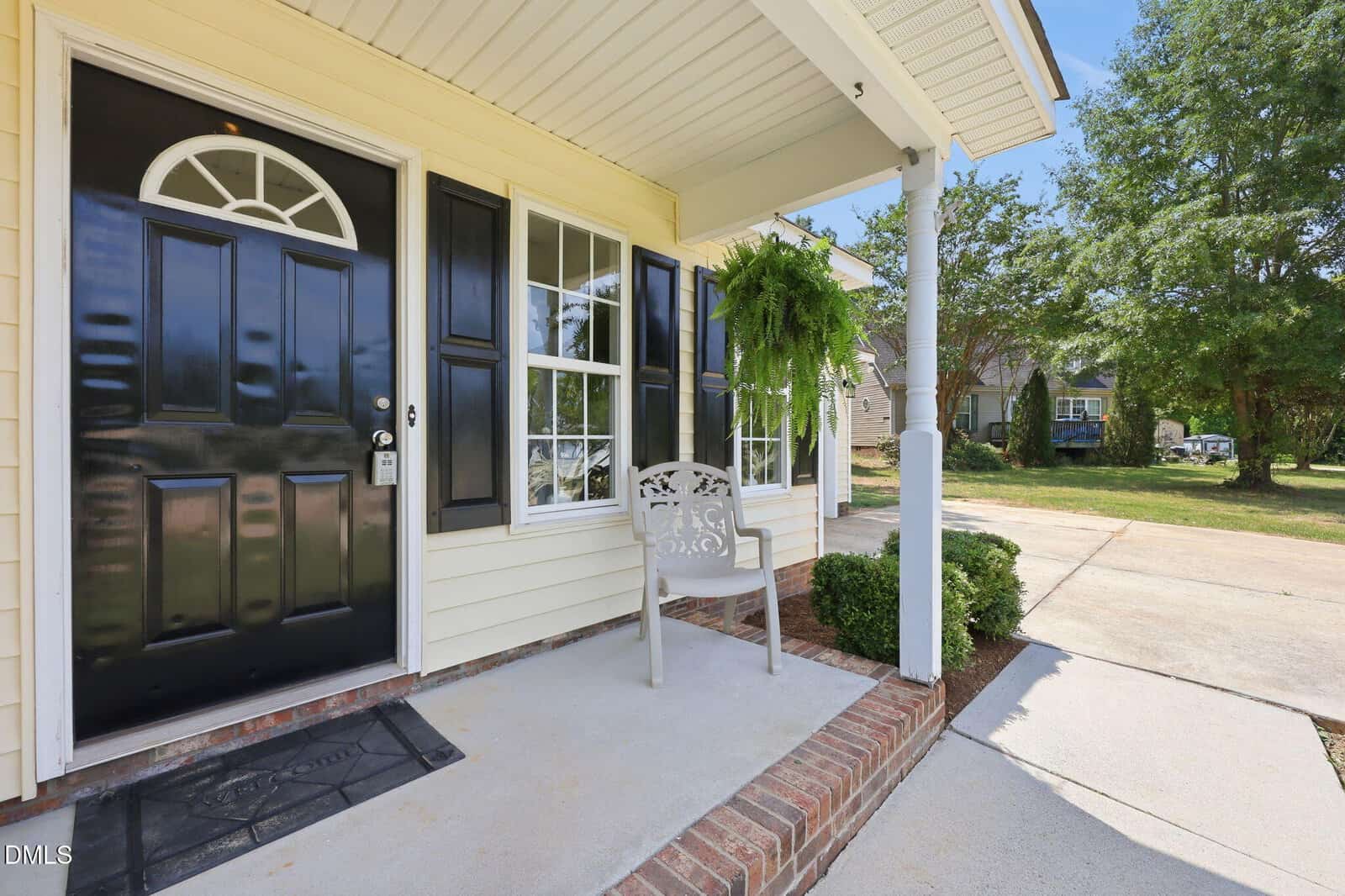 A covered front porch with a black door, decorative metal chair, hanging fern plant, and a concrete driveway. The lawn and trees are visible in the background under a clear blue sky.