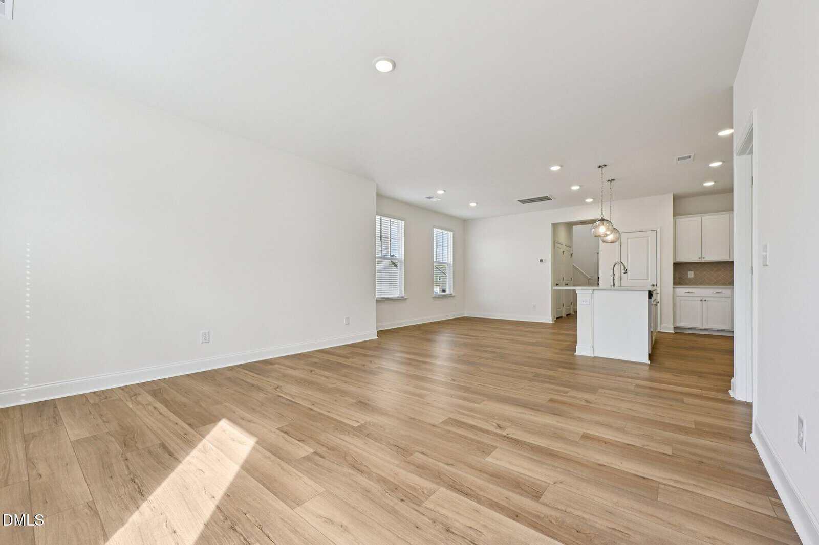 Bright, empty room with light wood floors, white walls, and an open kitchen area featuring white cabinets, an island, and pendant lighting. Multiple windows let in natural light.