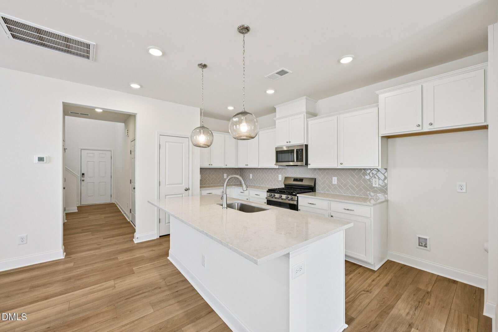 Modern kitchen with white cabinets, stainless steel appliances, a center island with sink, pendant lights, light wood flooring, and a tile backsplash. Staircase and entryway visible in the background.