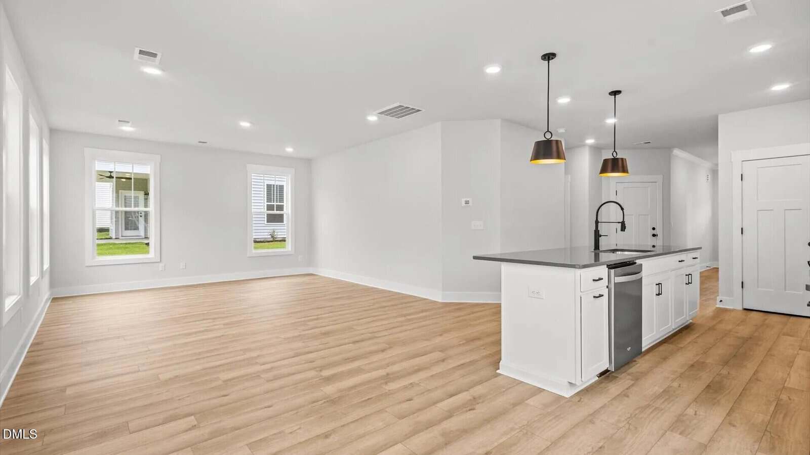Open, modern kitchen with island, pendant lights, stainless steel appliances, and light wood floors; adjacent to a bright living area with large windows and white walls.