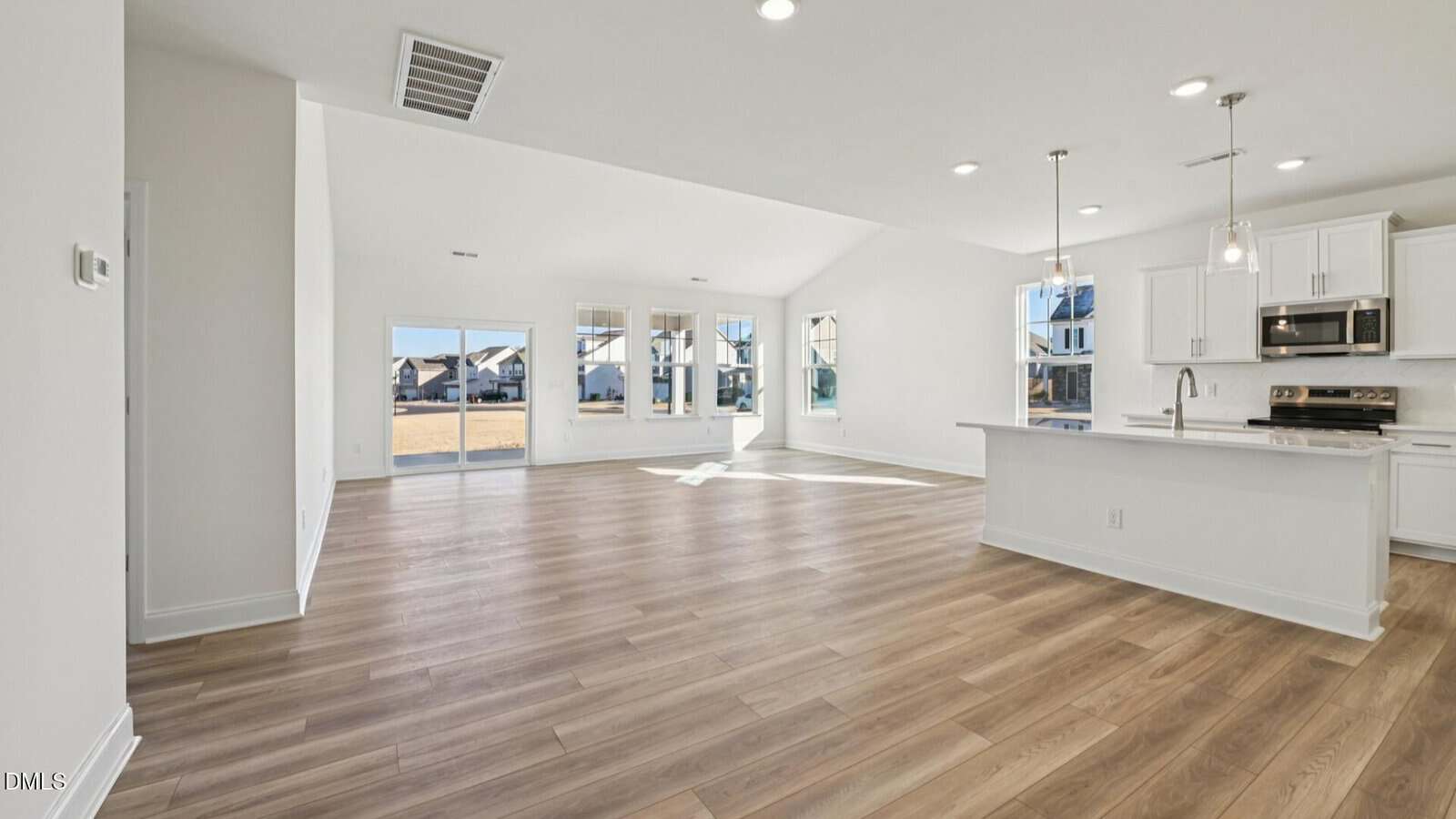 Bright, modern open-concept living space with wood floors, white walls, large windows, a kitchen with white cabinets, island, pendant lights, and stainless steel appliances. Sliding doors open to a backyard.