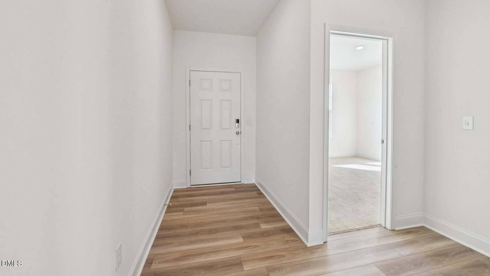 A hallway with light-colored walls and wood flooring leads to a white front door; an open doorway on the right reveals a carpeted room with sunlight coming in.