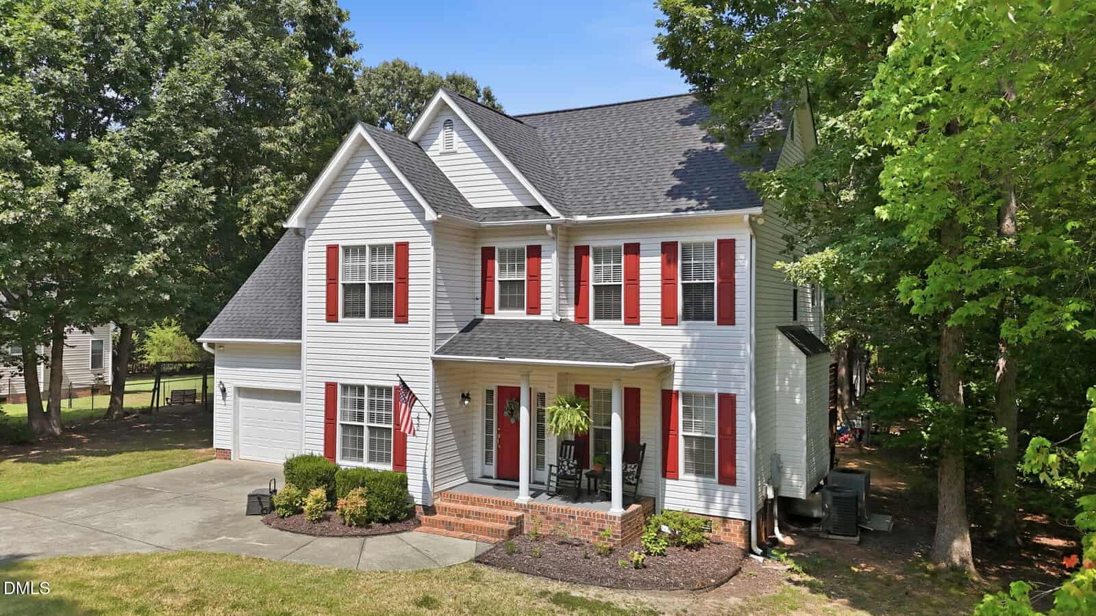 Two-story white house with red shutters and black roof, front porch with chairs and potted plant, American flag, driveway, green lawn, and surrounded by mature trees on a sunny day.