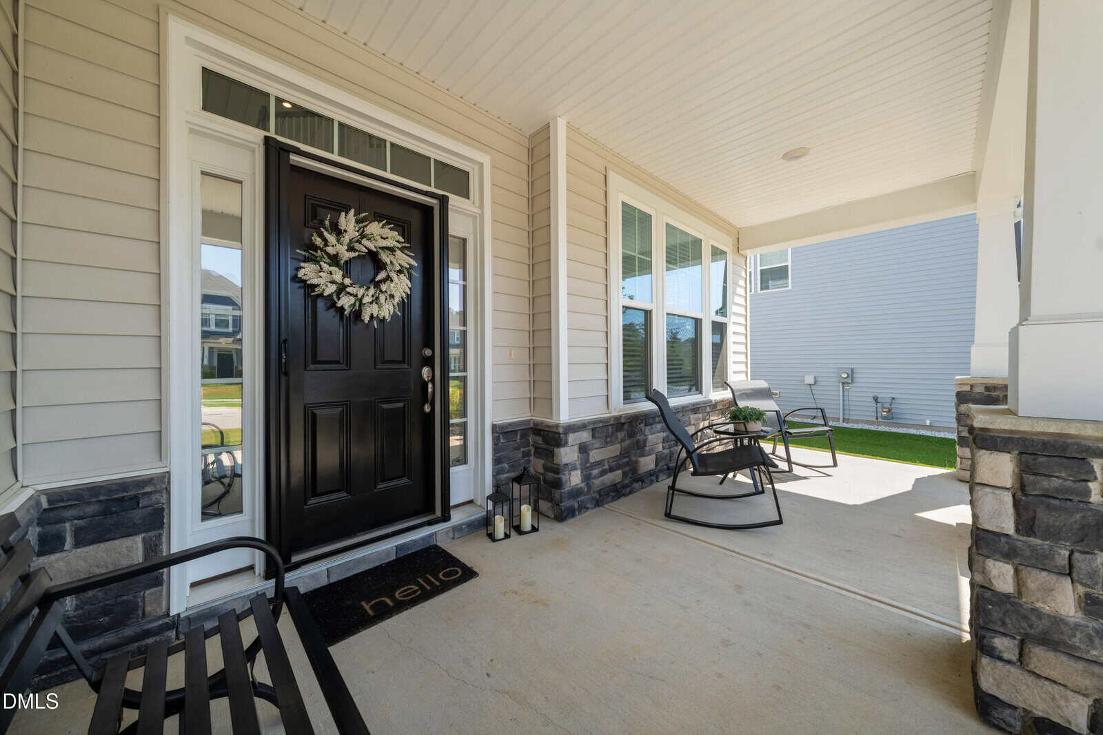 Covered porch with a black front door decorated with a wreath, rocking chairs, side table, lanterns, a bench, and a doormat that says “hello.” Stone and siding accents line the porch area.