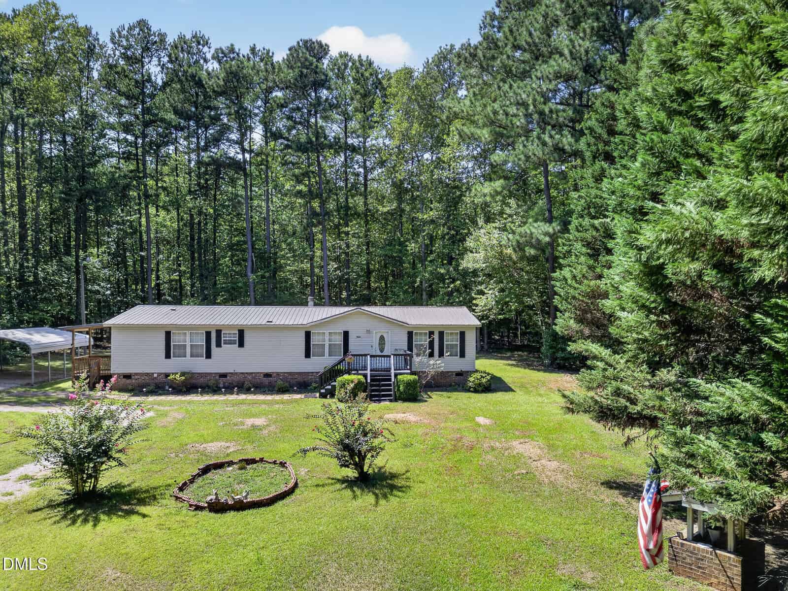 Charming single-story white house with a black roof, front steps, and a small garden, nestled at 7026 Southern Court in Oxford. Surrounded by tall trees and greenery, this home features an American flag on the right and a circular flower bed in the front yard.