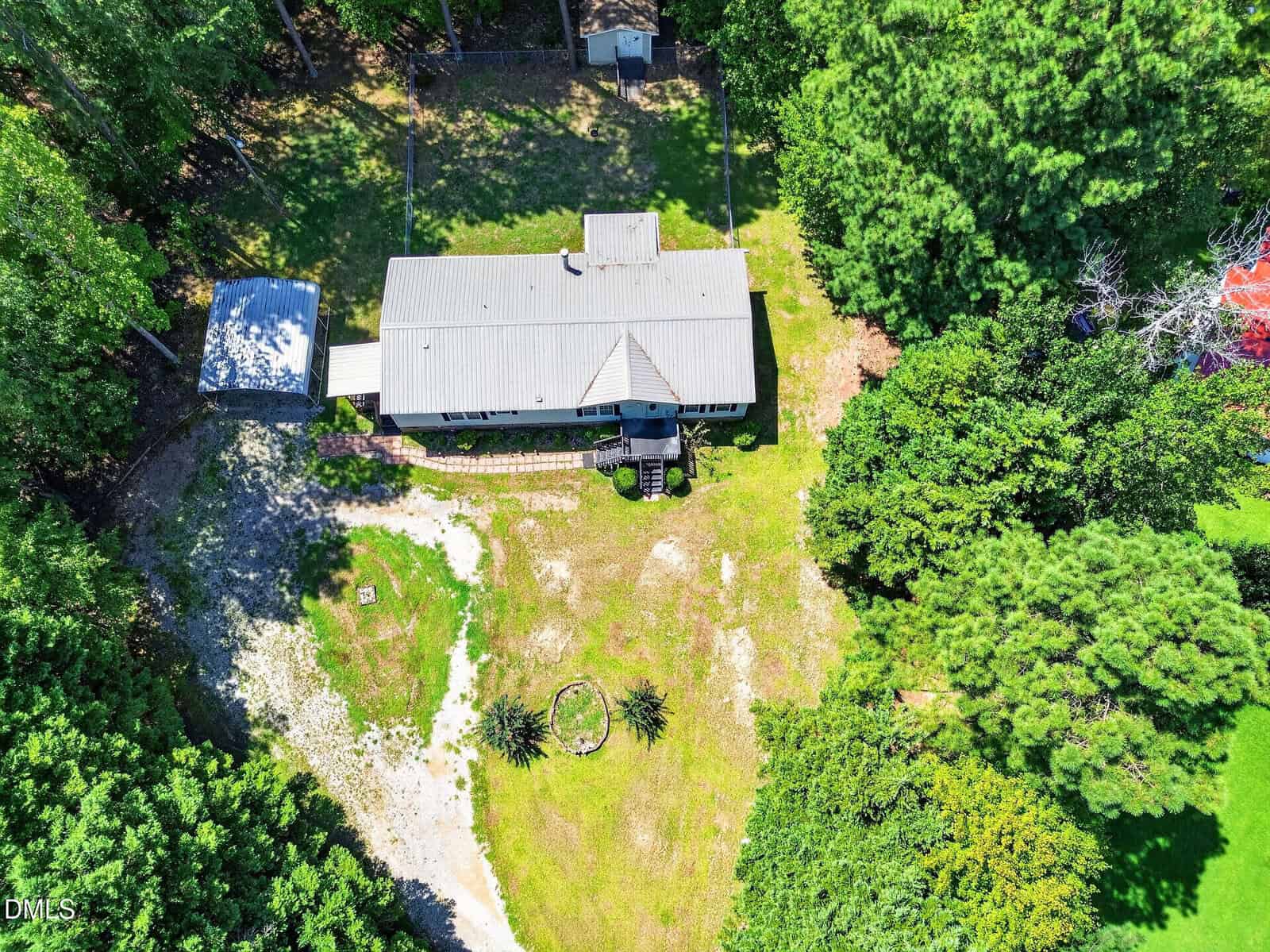Aerial view of a single-story house with a metal roof, surrounded by trees and a grassy yard. A circular fire pit and small garden are visible in the backyard, along with a gravel driveway.