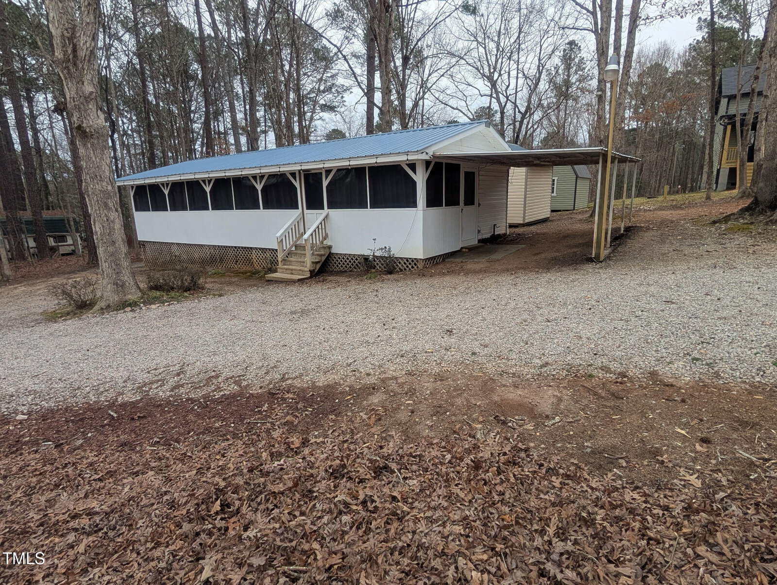 A white, screened-in house with a metal roof and front steps sits among trees at 122 Tomahawk Drive, Louisburg, next to a gravel driveway and a covered carport. Dry leaves and bare trees suggest it is autumn or winter.