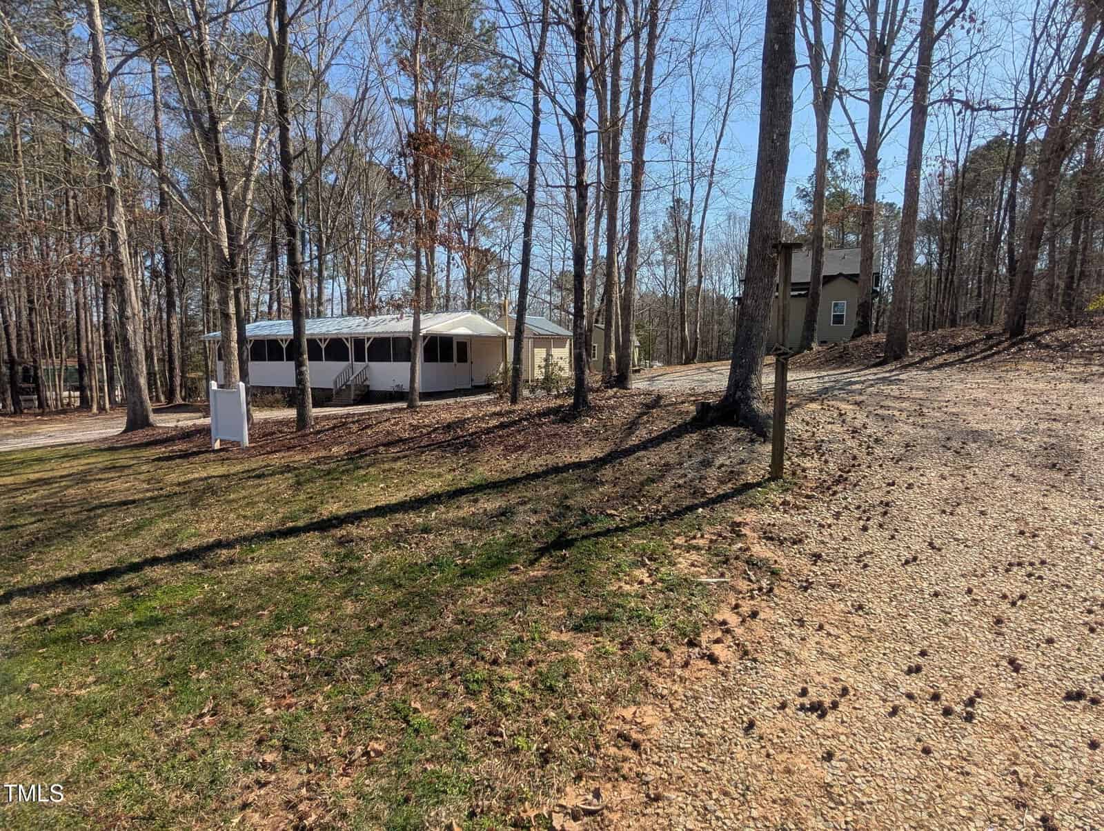 A gravel driveway leads to a small white house with a covered porch, surrounded by tall trees and scattered leaves on the ground. Another house is partially visible in the background.