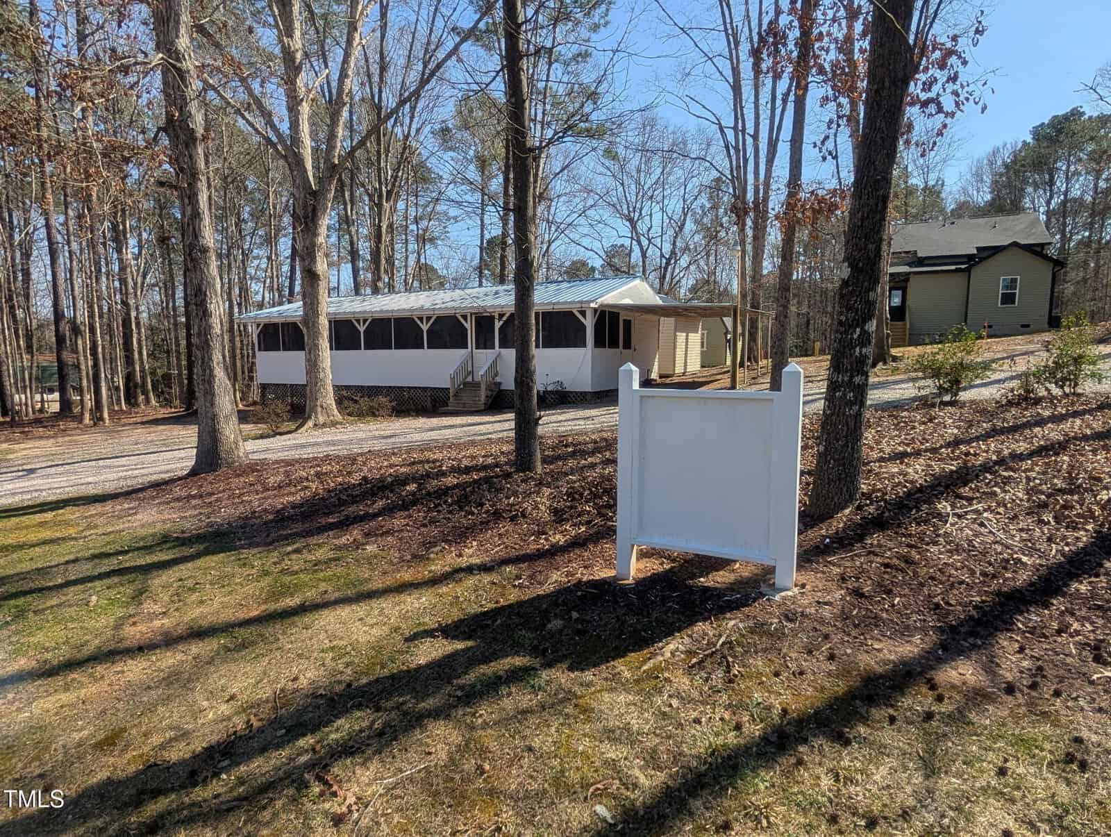 A white signpost stands in a wooded yard near a beige house with a screened-in porch; another house is visible in the background among tall trees under a clear blue sky.