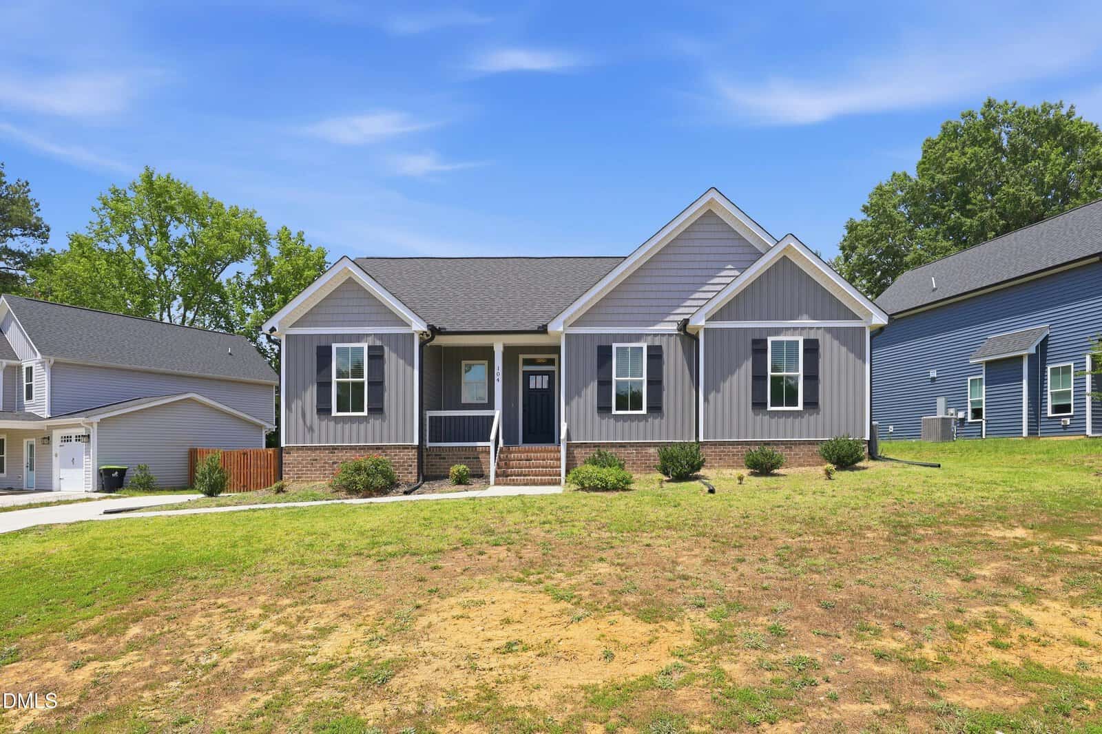 Single-story gray house with white trim, a small front porch, steps leading up to the door, and a patchy lawn under a blue sky with light clouds. Neighboring homes are visible on both sides.
